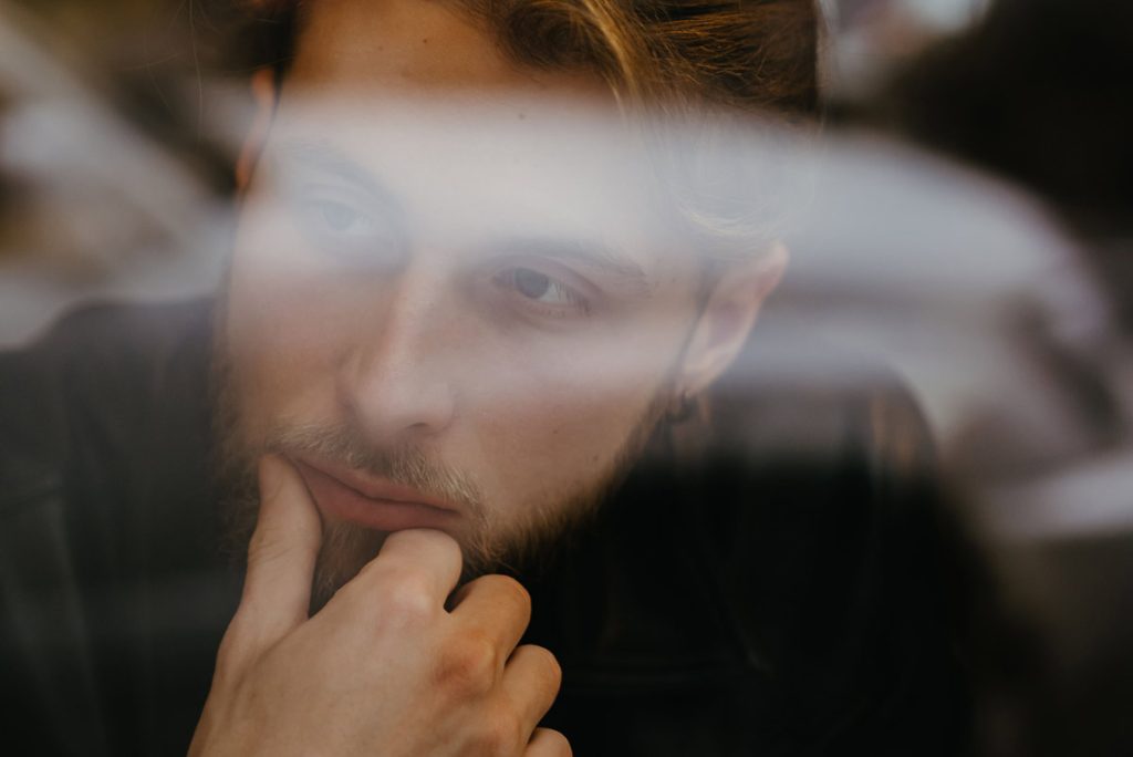 Young man with a beard rests his chin on his hand, looking thoughtfully through a window with reflections in the glass