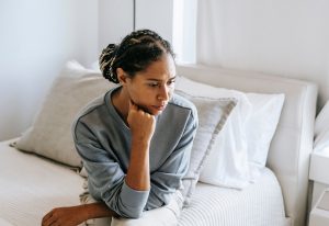 Woman sitting on a couch alone, resting her chin on her hand, looking worried and lost in thought.