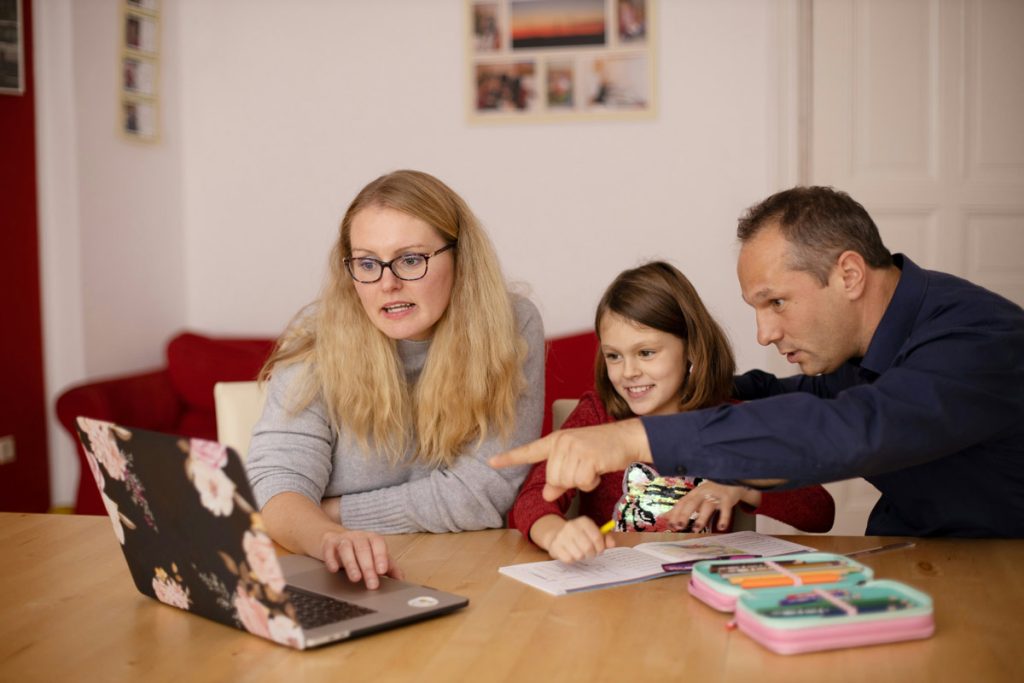 Two parents and their daughter looking at a laptop and homework together at the kitchen table.