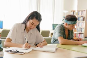 Two college students studying at tables in a campus library, one writing in a notebook and the other reading.