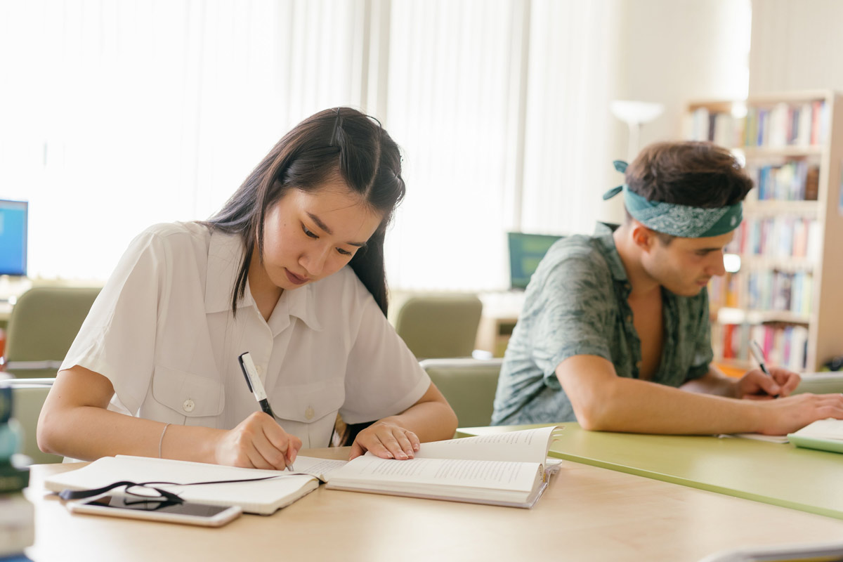 Two college students studying at tables in a campus library, one writing in a notebook and the other reading.