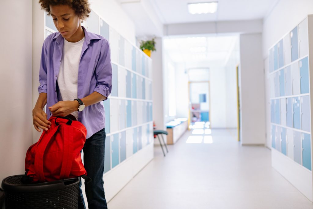 Teen boy packing a red duffel bag in a school hallway near lockers.