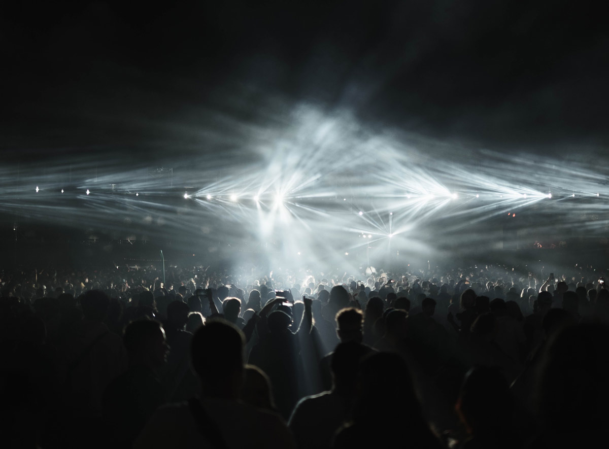: Silhouetted crowd at a nighttime concert or festival under dramatic white stage lighting.