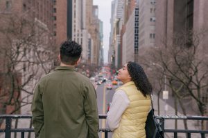 A young man and woman look out over a busy city street from an overlook, one glancing up with a calm expression.