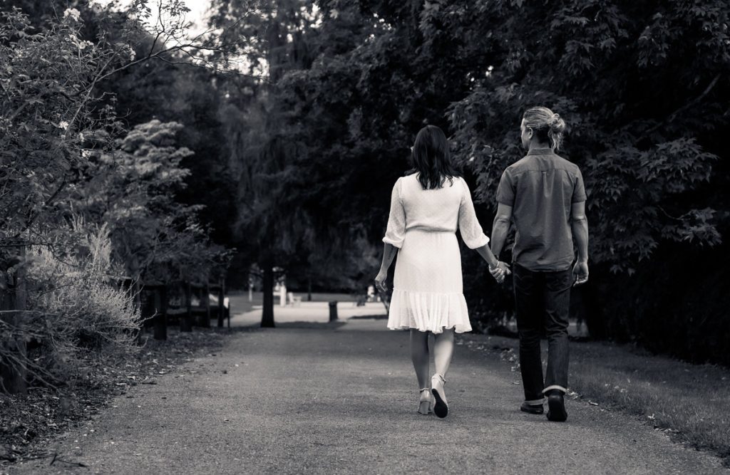 A couple walks hand in hand down a tree-lined park path, seen from behind in black and white.