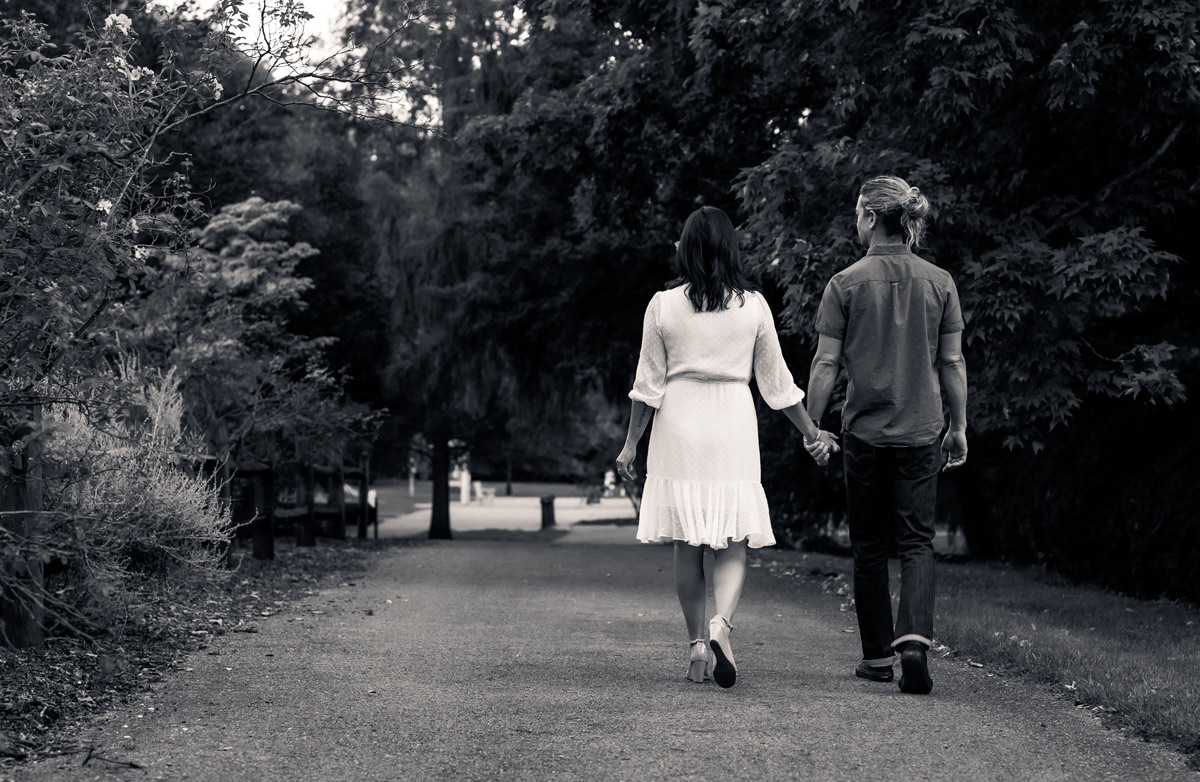 A couple walks hand in hand down a tree-lined park path, seen from behind in black and white.