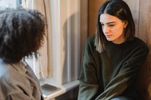 Young woman in a dark green sweater sits by a window, listening quietly to someone seated across from her.